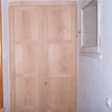 Une grande porte ou armoire en bois clair à panneaux est installée dans une pièce aux murs blancs, avec une petite fenêtre à droite.