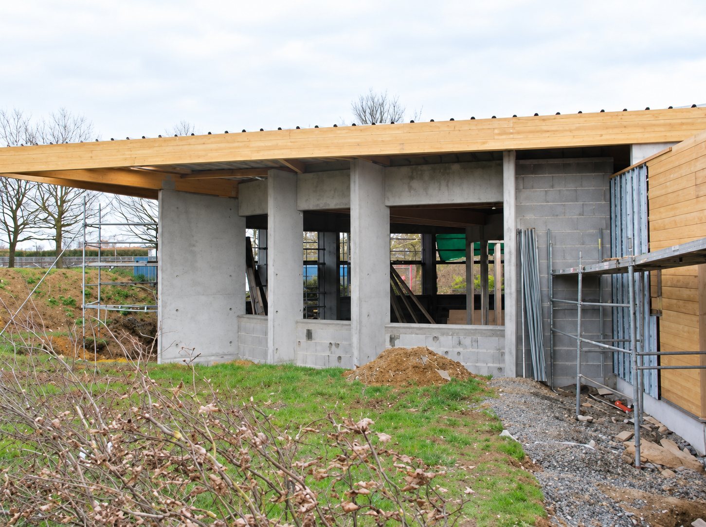 Chantier d'un bâtiment en blocs de béton brut avec une large toiture en bois en cours de construction, entouré d'échafaudages et de terre.