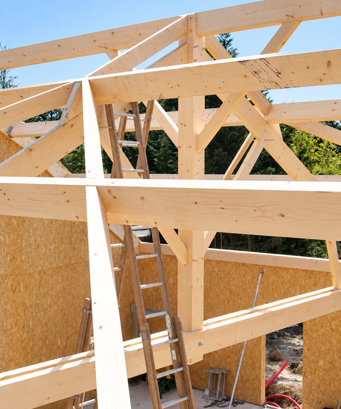 Charpente en bois clair en cours de montage sur une maison à ossature, avec des échelles appuyées contre les poutres et des panneaux OSB en arrière-plan.