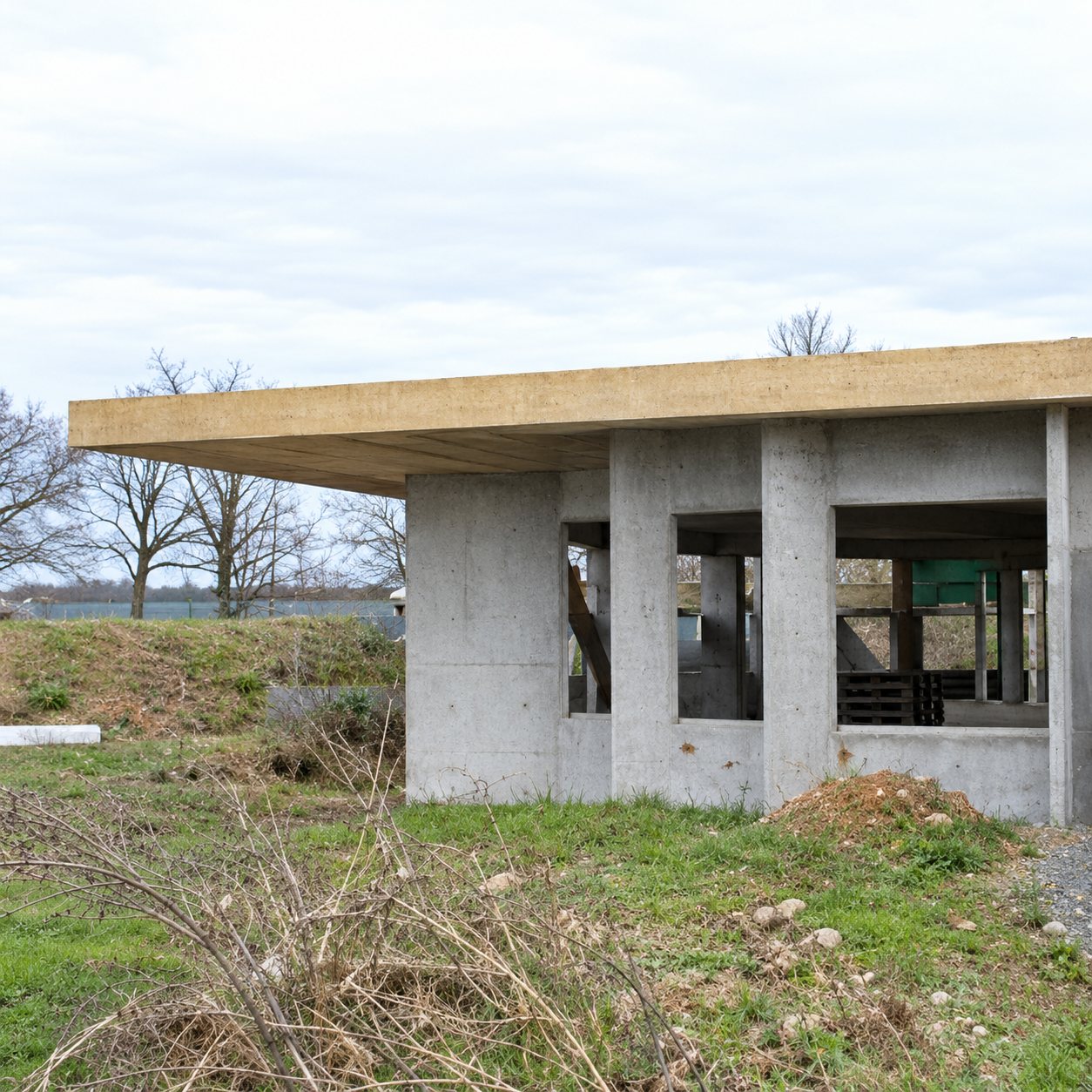 Un petit bâtiment en béton à toit plat débordant, entouré d'herbe et d'arbres nus, photographié sous un ciel gris.