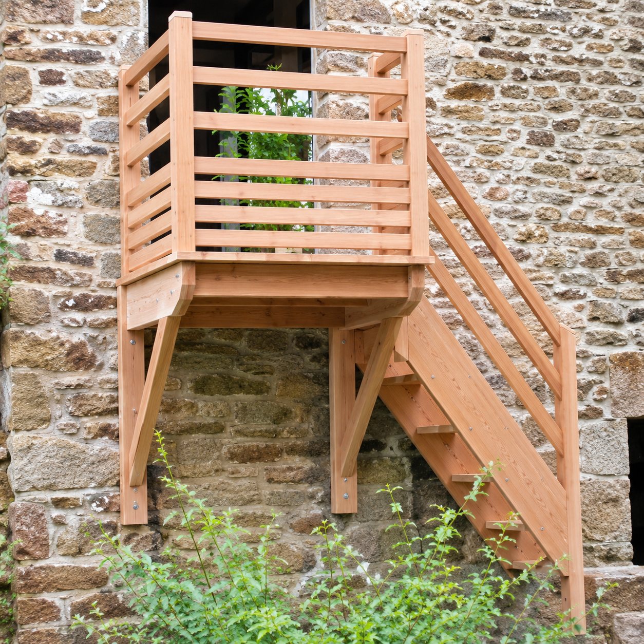 Un escalier extérieur en bois clair avec garde-corps et palier est fixé contre un vieux mur en pierre, avec un peu de verdure au premier plan.