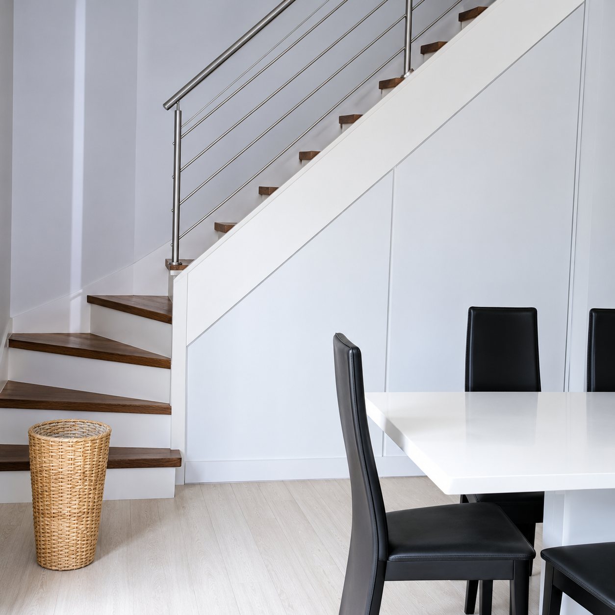 Un escalier blanc à marches sombres et garde-corps à câbles métalliques, près d'une table blanche entourée de chaises noires.