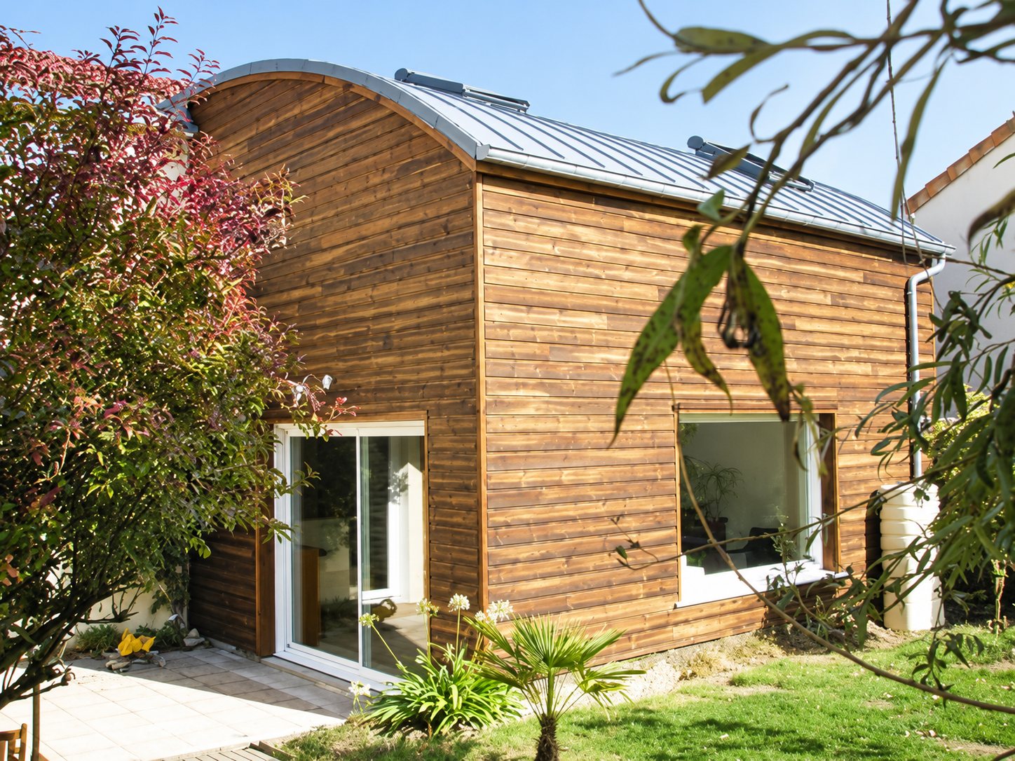 Une extension de maison en bardage bois avec un toit arrondi en zinc, une porte-fenêtre blanche et une grande fenêtre, entourée d'un jardin et de feuillages.