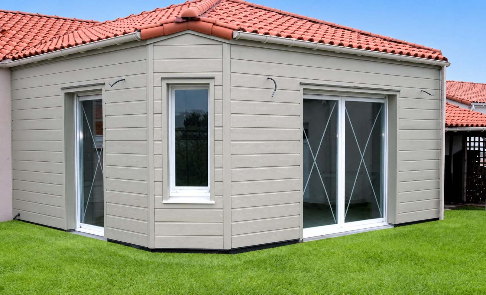 Extension de maison récente au bardage gris clair, dotée de baies vitrées coulissantes et d'une fenêtre blanche, sous un toit de tuiles rouges, au milieu d'une pelouse.