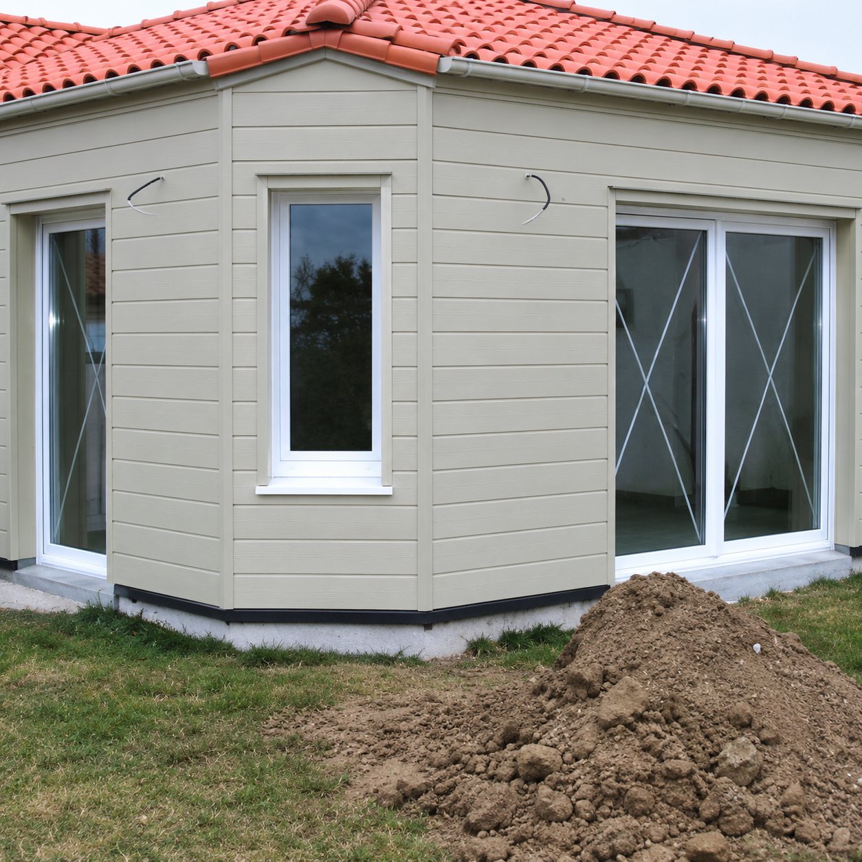 Une construction récente de forme polygonale aux murs clairs, couverte de tuiles orange, avec des fenêtres et une baie vitrée, et un tas de terre au premier plan.