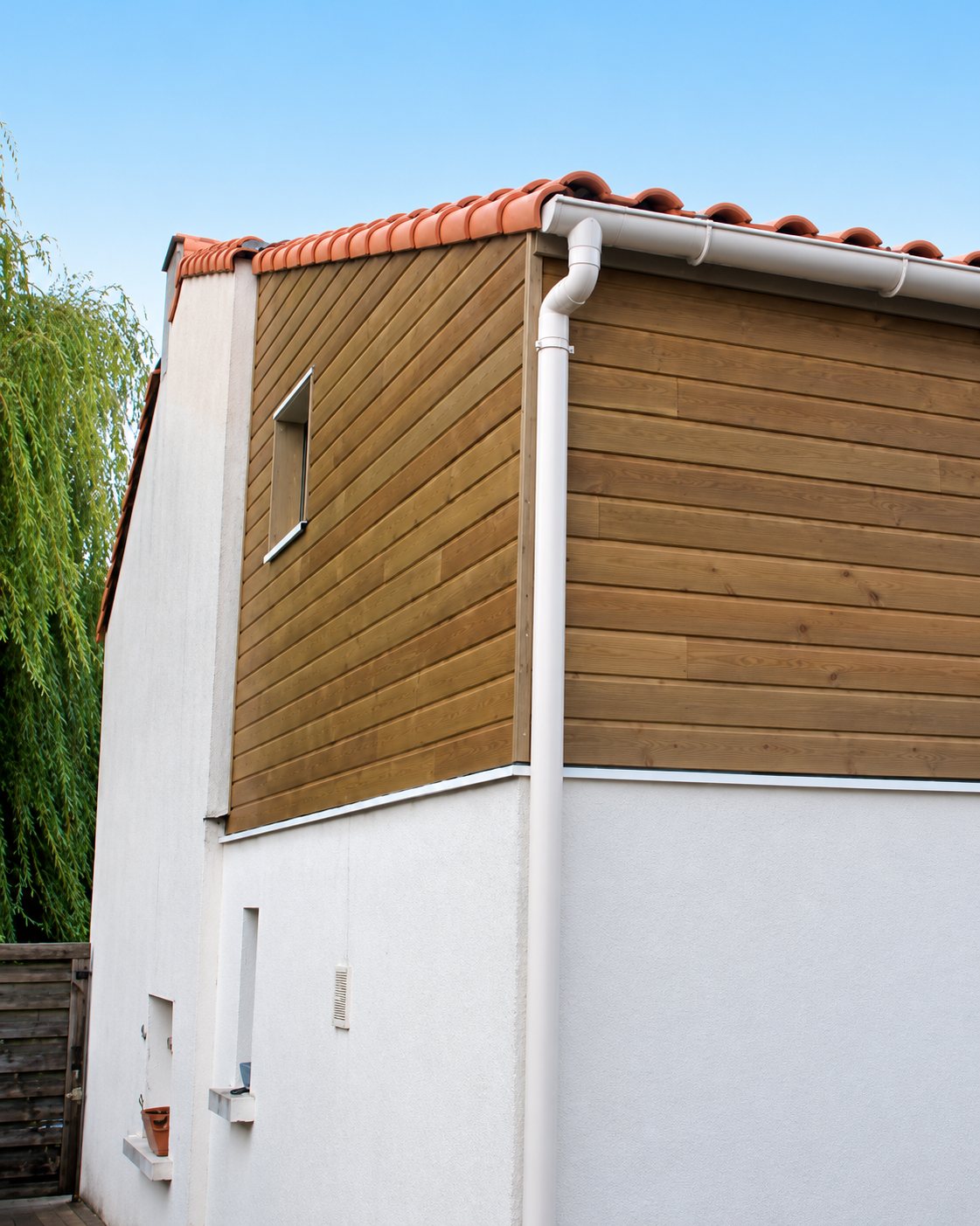 Angle d'une maison dont l'étage est habillé d'un bardage en bois, avec un rez-de-chaussée enduit en blanc, une gouttière blanche et un toit en tuiles rouges sous un ciel bleu.