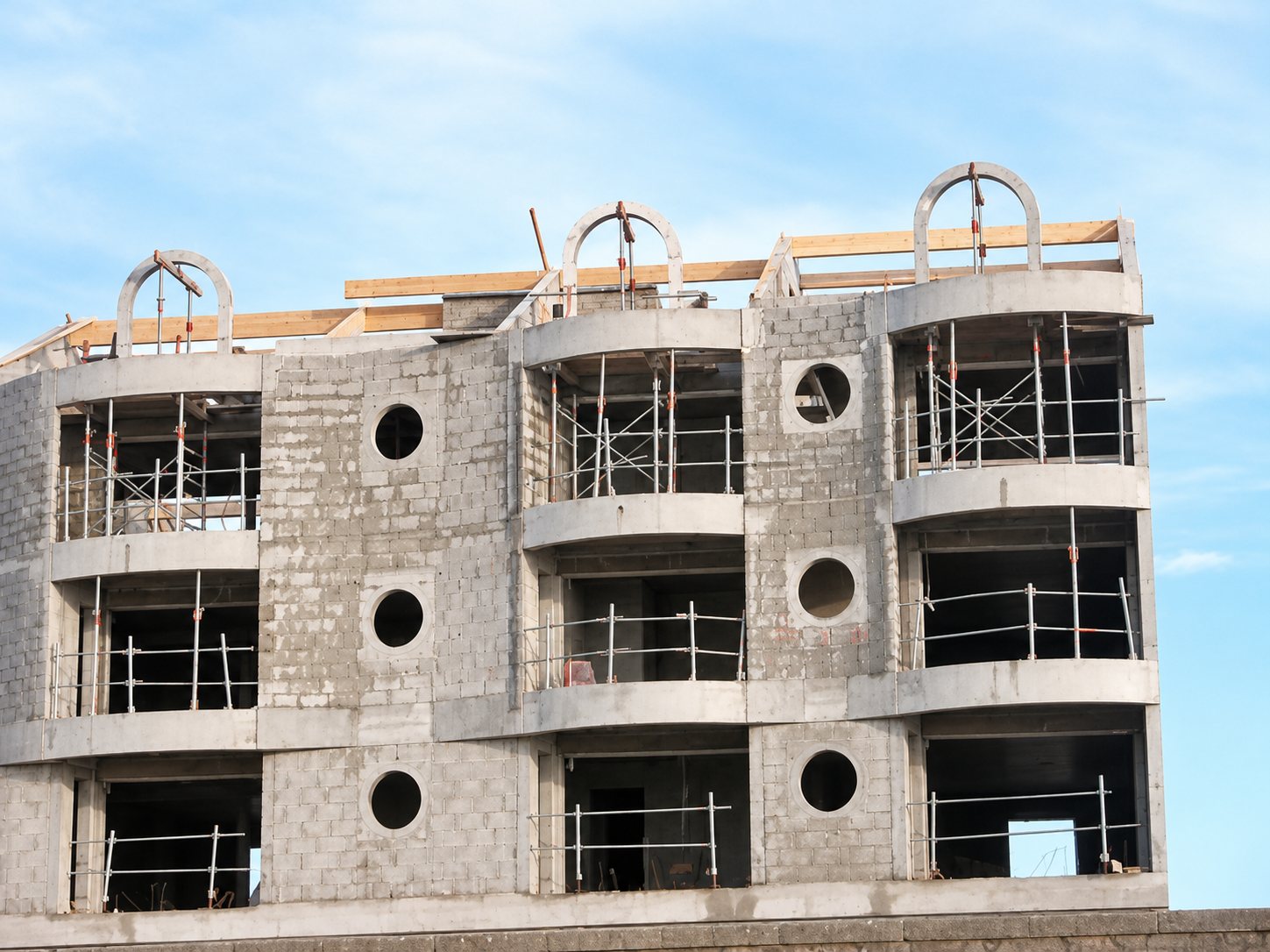 Façade d'un immeuble de quatre étages en construction, avec balcons courbes, échafaudages, fenêtres rondes et arches en métal sur le toit, sous un ciel clair.