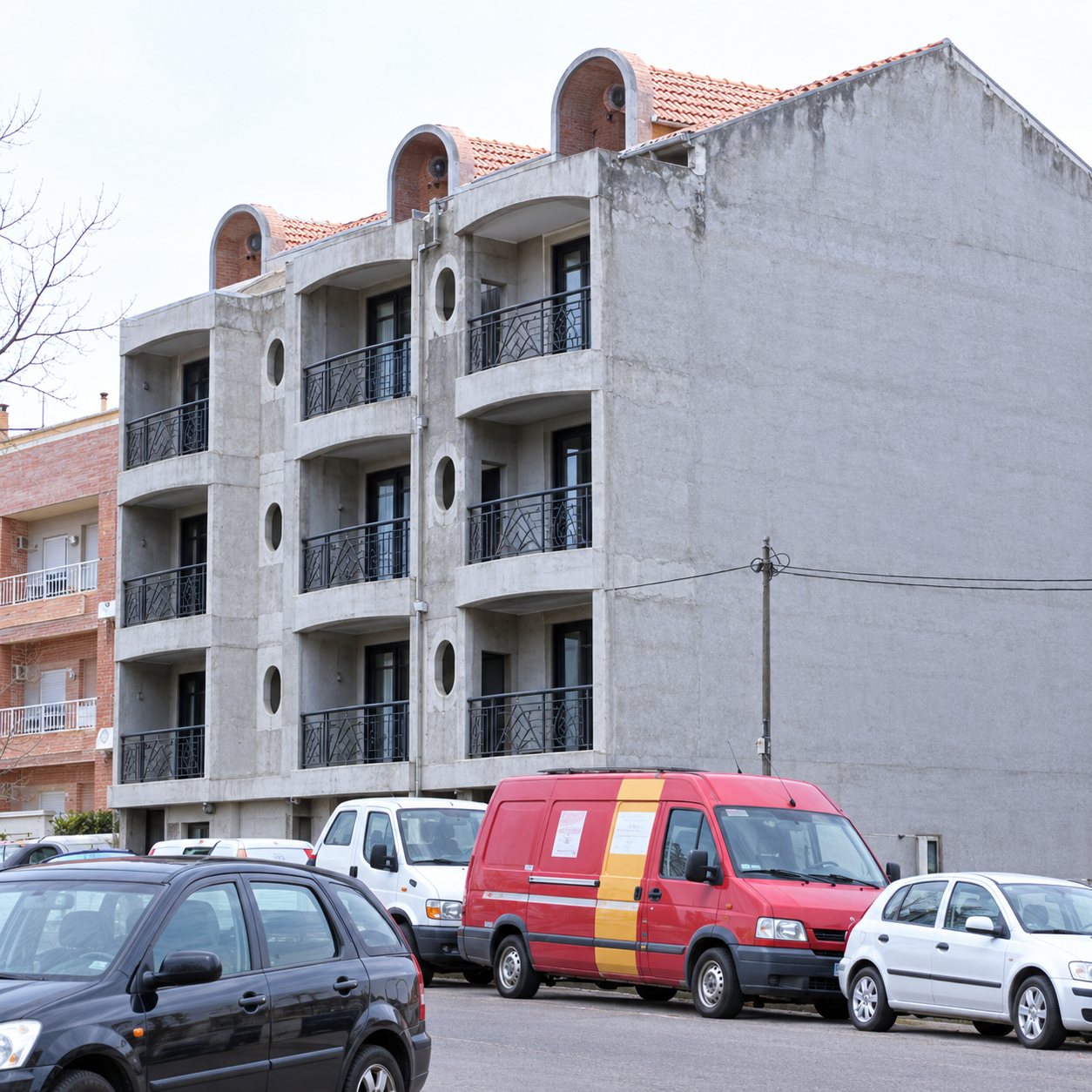 Un immeuble d'habitation en cours de construction à la façade grise, avec des balcons non terminés et plusieurs voitures et une camionnette rouge garées devant.