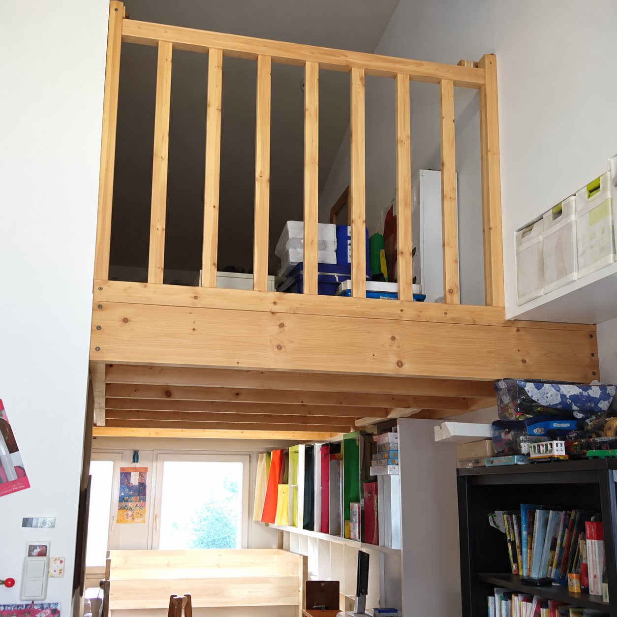 Une mezzanine en bois clair avec garde-corps à barreaux surplombe une pièce aménagée avec une étagère sombre.