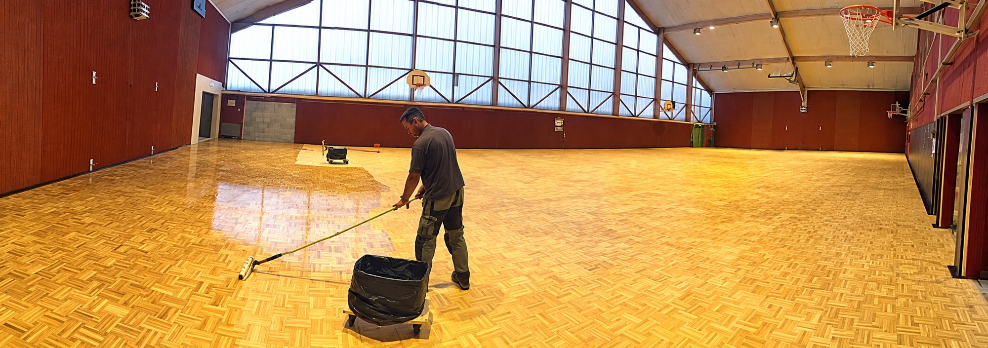 Un ouvrier étale du produit avec une raclette sur le parquet brillant d'une grande salle de sport équipée de paniers de basket.
