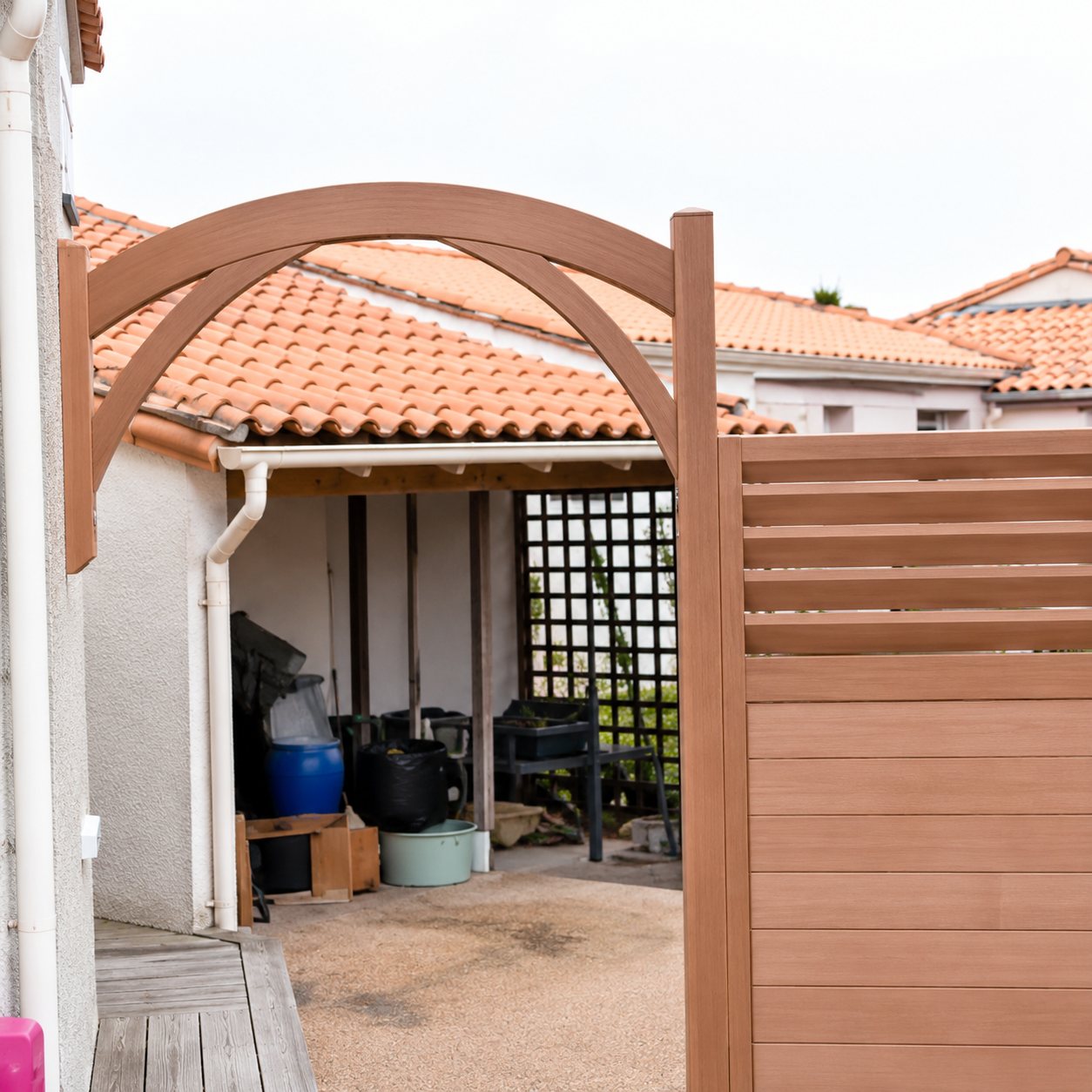 Une arche de pergola en bois clair et un panneau claustra se dressent devant une terrasse couverte de tuiles orange.