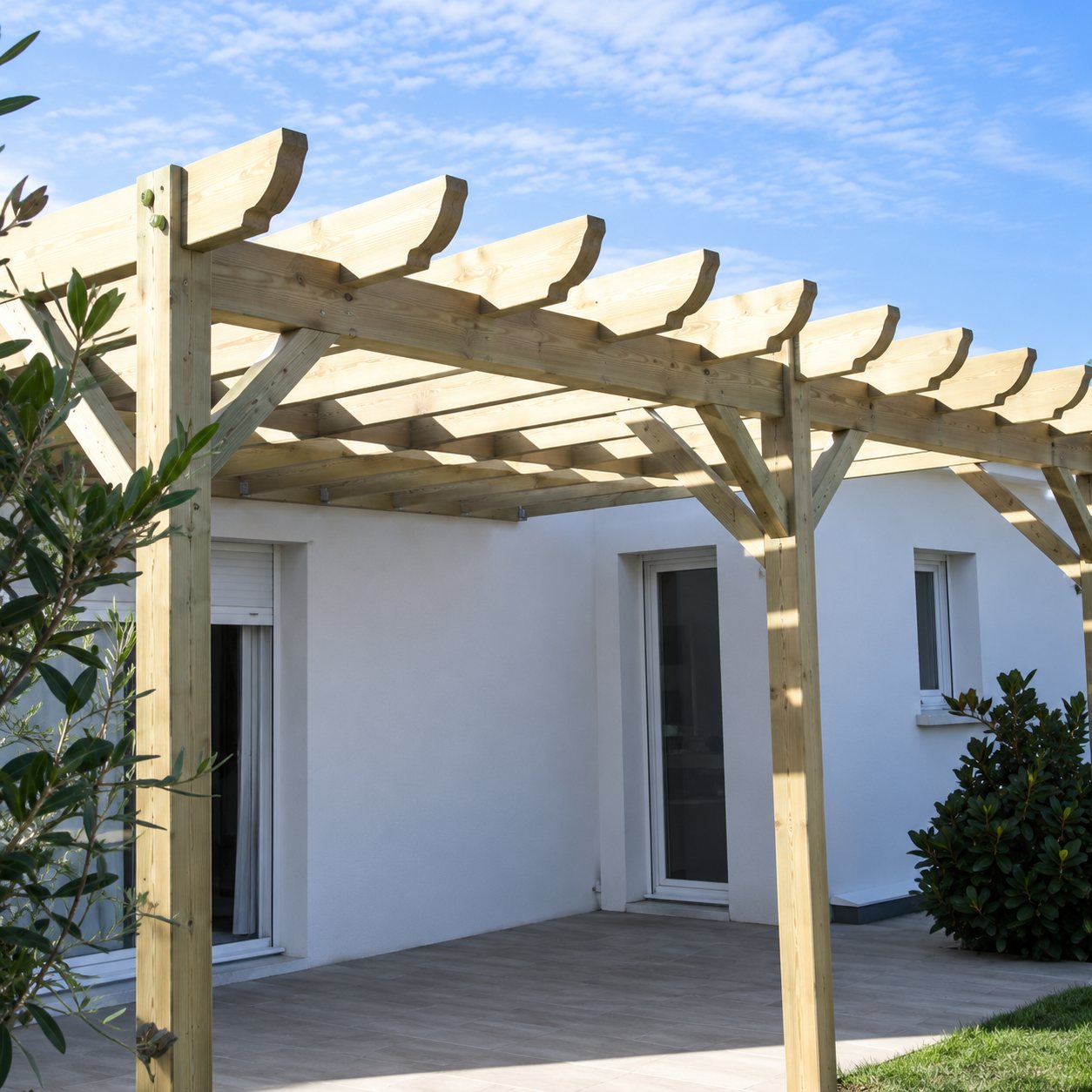 Une pergola en bois aux poutres arrondies est installée contre une maison blanche, sous un ciel bleu dégagé.