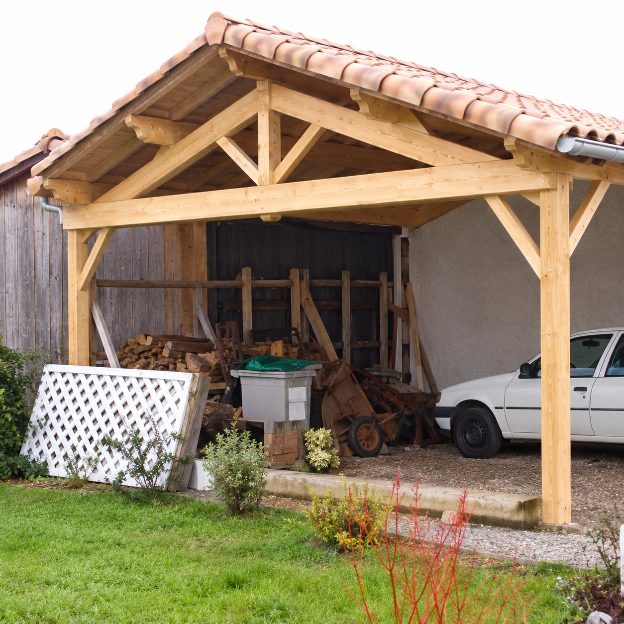 Une pergola en bois clair au toit de chaume est installée devant une maison, avec une voiture blanche garée à côté et de l'herbe au premier plan.