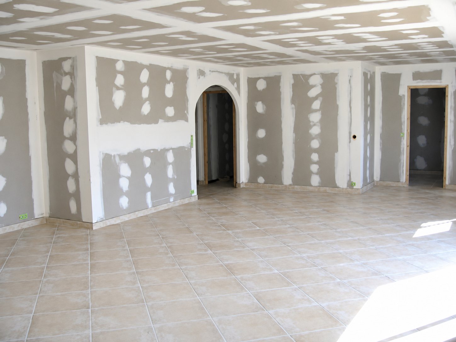 Une vaste pièce en cours de rénovation avec des murs en plaques de plâtre jointoyées, une arche, un plafond en placo et un sol récemment carrelé.