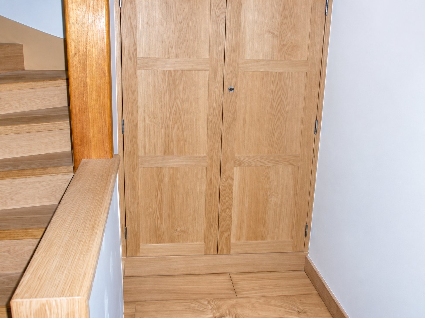 Placard à deux portes en chêne clair encastré dans un mur à côté d'un escalier avec rampe en bois et parquet au sol.