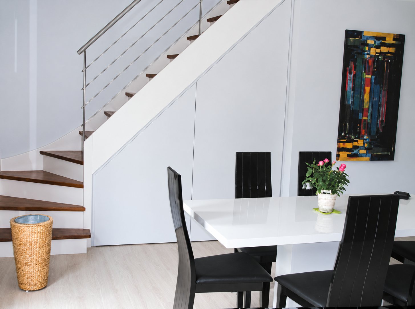 Salle à manger lumineuse avec une table blanche laquée, des chaises noires, un escalier à marches en bois et rambarde métallique, et un tableau coloré au mur.