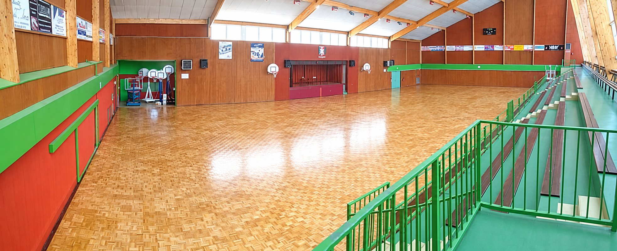 Vue panoramique d'une grande salle de sport au parquet clair, avec des murs en bois, rouges et verts et des gradins sur la droite.