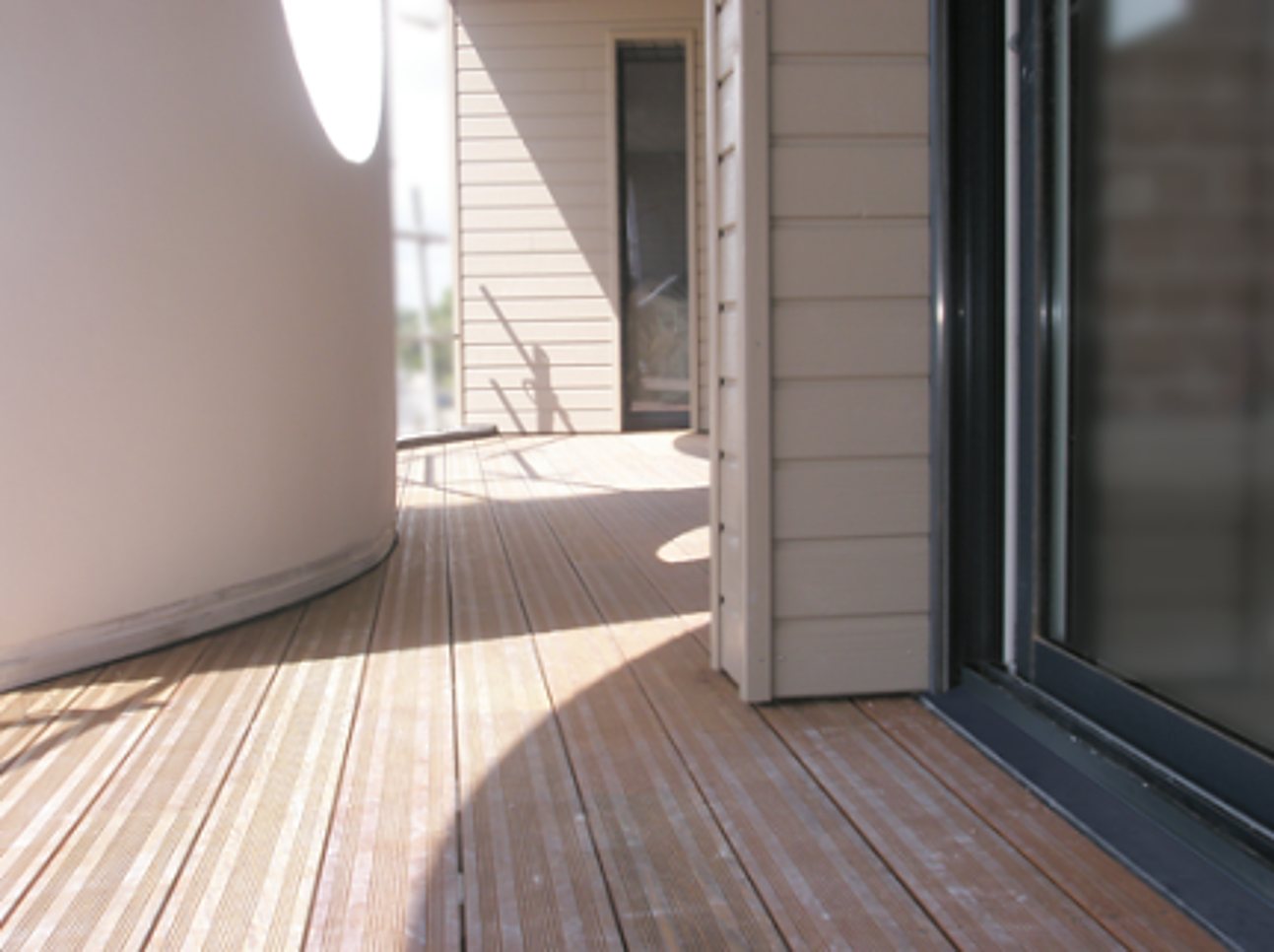 Une terrasse en lames de bois longe un mur incurvé et un bardage clair, près d'une grande baie vitrée, en plein soleil.