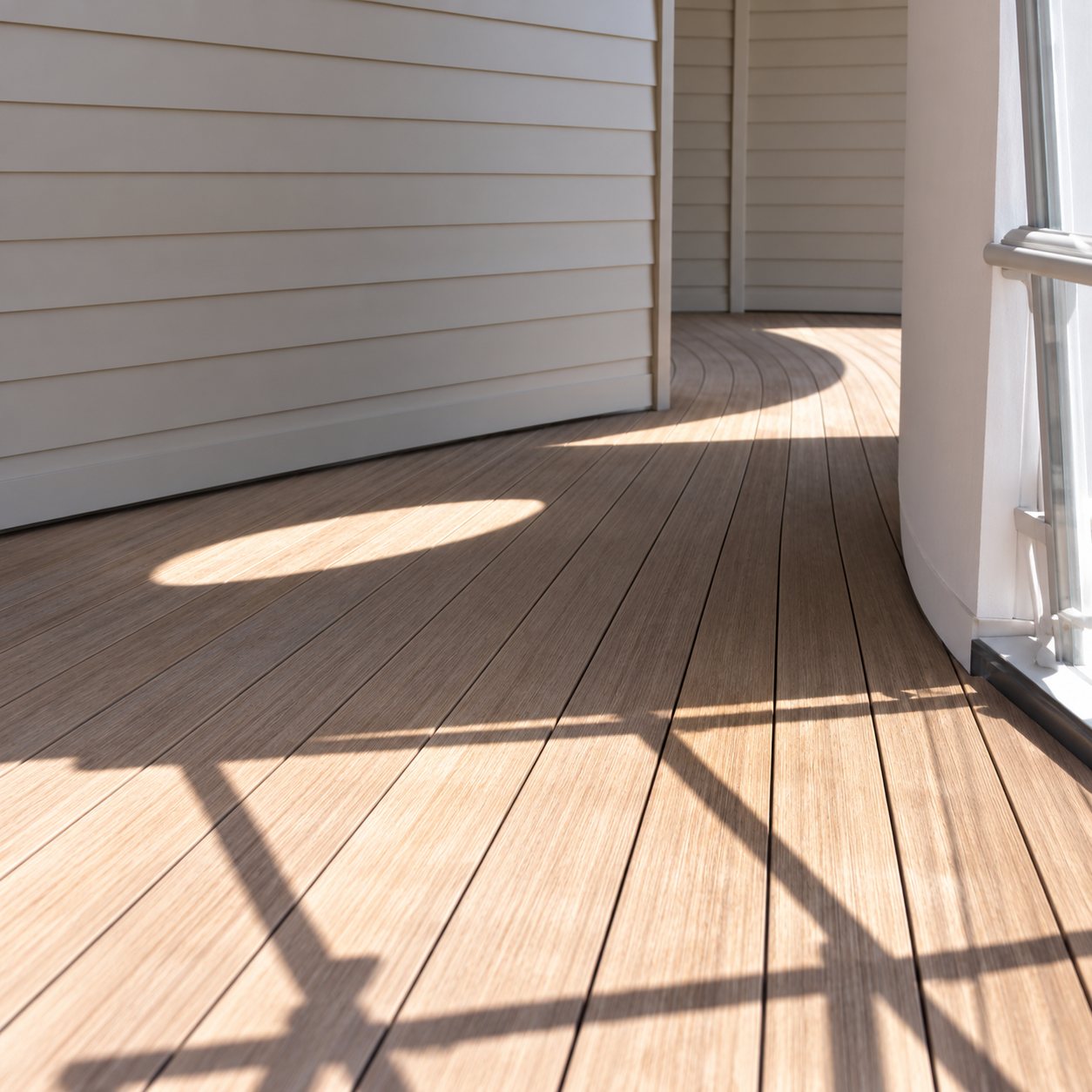 Une terrasse en lames de bois baignée de soleil, avec des ombres de barreaux projetées au sol le long d'un mur clair.