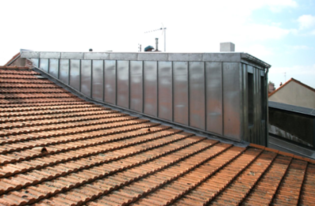 Vue d'un toit en tuiles rouges canal avec une grande lucarne recouverte de panneaux de zinc à joints debout sous un ciel clair.